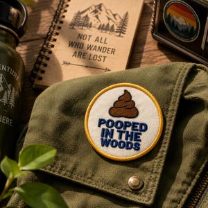 May include: A circular embroidered patch featuring a brown poop emoji icon above the blue text "POOPED IN THE WOODS" on a white background with a yellow border, attached to an olive green fabric surface with a metal snap button closure.
