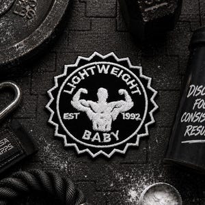 Può includere: Una toppa ricamata circolare con un bordo seghettato a forma di stella. La toppa presenta uno sfondo nero con ricami bianchi che raffigurano una silhouette muscolosa che flette le braccia, il testo LIGHTWEIGHT BABY e il testo EST 1992.