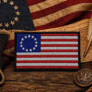 Puede incluir: Un parche patriótico con la bandera de Betsy Ross, con un cantón azul con 13 estrellas y rayas rojas y blancas. El parche se presenta sobre un fondo de una bandera estadounidense, una pluma y un botón vintage, evocando la historia y el patriotismo americano.