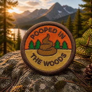 Peut inclure: Écusson rond brodé avec le texte "POOPED IN THE WOODS". L'écusson présente un emoji caca marron, des arbres verts et un fond orange et jaune. L'écusson est placé sur un fond de montagne pittoresque.