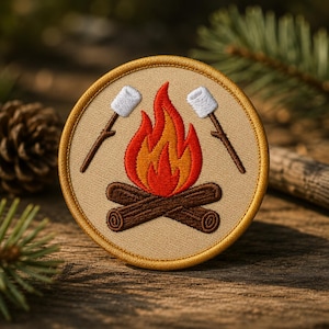 Könnte beinhalten: Ein runder bestickter Aufnäher mit einem Lagerfeuer-Motiv auf beigem Hintergrund. Der Aufnäher zeigt zwei braune Holzscheite an der Basis, eine leuchtend orange und rote Flamme in der Mitte sowie zwei weiße Marshmallows auf braunen Stöcken.