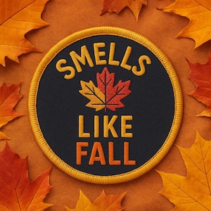 Puede incluir: Parche circular con la frase "SMELLS LIKE FALL" en hilo dorado. Presenta una hoja de arce naranja y roja sobre fondo negro, bordeada en dorado. El fondo es naranja con hojas de otoño.