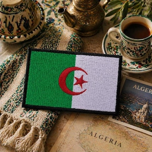 May include: A rectangular patch featuring the flag of Algeria. The flag is divided vertically into green and white sections, with a red crescent and star emblem in the center. The patch is bordered in black and rests on a patterned surface.