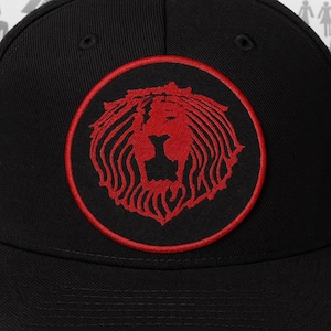 May include: Black baseball cap featuring a red lion's head emblem within a red circular border. The lion's head is intricately detailed, and the cap has a curved brim and a structured crown.