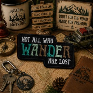 May include: A black patch with the text "NOT ALL WHO WANDER ARE LOST" in white, green, and brown embroidery. The image includes a compass, a mug with "FUELED BY ADVENTURE", a miniature SUV, and a sign that says "BUILT FOR THE ROAD MADE FOR FREEDOM".