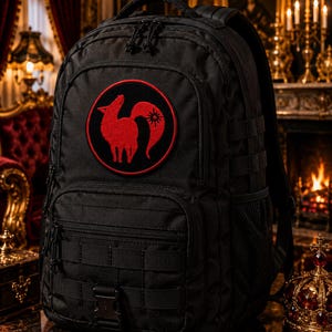 Pode incluir: Uma mochila t&aacute;tica preta com um emblema circular vermelho no bolso frontal. O emblema exibe a silhueta estilizada de uma raposa vermelha com um s&iacute;mbolo solar na cauda. A mochila inclui v&aacute;rios compartimentos com z&iacute;per e tiras de fixa&ccedil;&atilde;o.