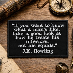 Könnte beinhalten: Ein rechteckiger, schwarzer, bestickter Aufnäher mit weißem Text: "If you want to know what a man's like, take a good look at how he treats his inferiors, not his equals." Der Name J.K. Rowling steht am unteren Rand des Aufnähers.