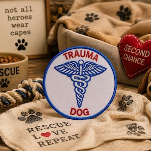 May include: A circular embroidered patch with a white background and a blue border. It features a blue caduceus symbol in the center with the red text TRAUMA above and DOG below. The patch is displayed among various pet-themed items.