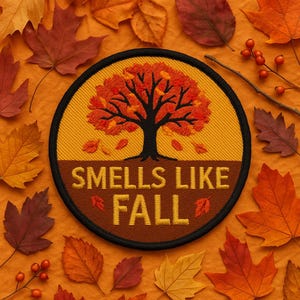 Puede incluir: Parche bordado con temática otoñal. El parche presenta un árbol con hojas naranjas y rojas, y el texto "SMELLS LIKE FALL" en letras doradas. El parche está rodeado de hojas de otoño en tonos naranja, rojo y marrón.