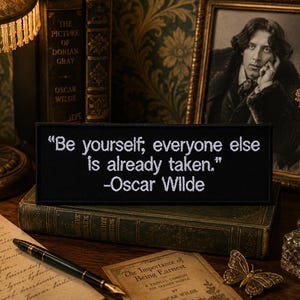 Puede incluir: Un parche rectangular negro bordado con el texto en blanco "Be yourself; everyone else is already taken." seguido de la atribución "-Oscar Wilde" en una fuente sans-serif clara, presentado como un artículo de tela plano.