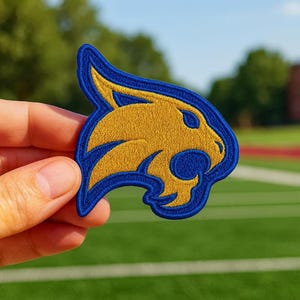 Puede incluir: Parche bordado con la cabeza estilizada de un lince en oro y azul. El parche tiene un diseño detallado con un borde azul, probablemente para ropa o accesorios. El parche se sostiene sobre un fondo verde.