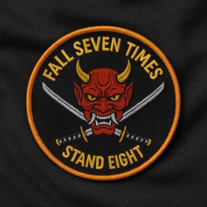 May include: Embroidered patch with a red demon face, yellow horns, and crossed swords. The patch has the text "FALL SEVEN TIMES STAND EIGHT" in yellow. The patch is round with a black background and an orange border.