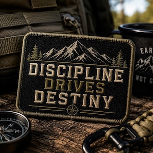 Puede incluir: Un parche rectangular negro bordado que presenta una cadena montañosa y pinos sobre el texto "DISCIPLINE DRIVES DESTINY" en letras color canela, con un pequeño icono de brújula centrado en el borde inferior y un borde cosido.