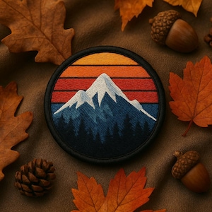 May include: Round embroidered patch featuring a mountain range with a sunset design. The patch has a black border, with a white mountain peak, blue forest, and orange and red sunset. The patch is surrounded by autumn leaves, acorns, and a pinecone.