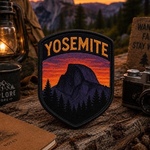 May include: Embroidered patch featuring the word "YOSEMITE" in gold lettering above a mountain scene at sunset. The patch has a black border and is set against a rustic wooden surface, with a vintage camera and camping lantern in the background.