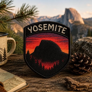 May include: A shield-shaped embroidered patch featuring the word YOSEMITE in white text at the top. The design depicts a dark silhouette of Half Dome against a vibrant red and orange sunset sky, with black pine tree silhouettes at the base.