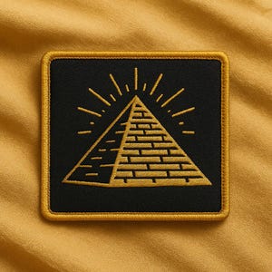 May include: Square black patch with a gold embroidered pyramid design. The pyramid is centered with radiating lines above it. The patch has a gold border and is set against a mustard yellow background.