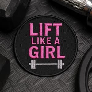 Puede incluir: Un parche redondo negro con la frase bordada en rosa "LIFT LIKE A GIRL". Un gráfico de barra con pesas está debajo del texto. El parche está rodeado por un borde negro y es probablemente un artículo decorativo.