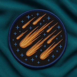 May include: Embroidered patch featuring a circular design with a black background and a blue border. The patch showcases several orange comet illustrations and blue star accents, creating a celestial theme. The patch is set against a teal fabric background.
