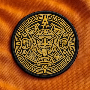 Peut inclure: Un écusson circulaire avec un motif de calendrier aztèque doré sur fond noir. L'écusson est posé sur un tissu orange texturé, mettant en valeur les motifs et symboles complexes du calendrier ancien.