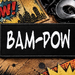 May include: A rectangular black embroidered patch featuring the white text BAM-POW in a jagged, comic book style font. The patch has a thick black stitched border and is centered horizontally, showcasing a bold, high-contrast design.