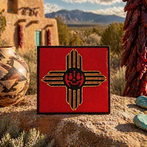 May include: A square, red patch featuring a stylized Zia sun symbol in black and gold. The patch is set against a backdrop of a Southwestern landscape, including a traditional adobe building and mountains.