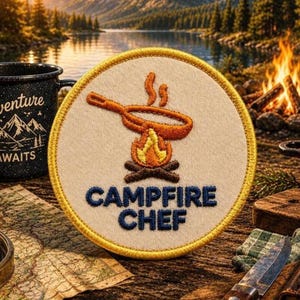 May include: A circular embroidered patch with a cream-colored background and a yellow border. The design features an orange frying pan over a campfire with flames and brown logs. The text CAMPFIRE CHEF is embroidered in dark blue letters.