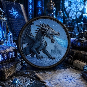 May include: A circular embroidered patch featuring a dark blue dragon with sharp scales and wings, exhaling a stream of light blue frost toward a small castle silhouette. The design is rendered in shades of blue and white with a black border.
