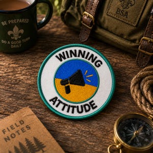 May include: A circular embroidered patch with a teal border, featuring a blue and yellow background, a black megaphone icon, and the black text "WINNING ATTITUDE" arched along the top and bottom edges of the white circular border.