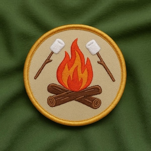 May include: Embroidered patch featuring a campfire scene. The design includes a stylized orange and yellow flame, two logs, and two marshmallows on sticks. The patch has a tan background with a gold border, set against a green fabric.