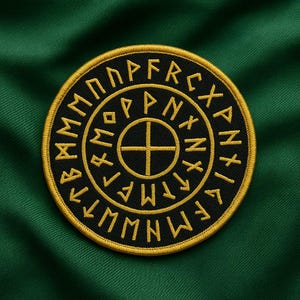 May include: A round, embroidered patch with a black center and gold-colored border and lettering. The patch features a cross design in the center and runes around the perimeter. The background is a dark green fabric.