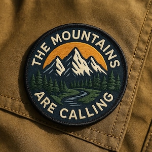 May include: A round patch with the words "THE MOUNTAINS ARE CALLING". The patch shows a mountain range, a setting sun, a river, and evergreen trees. The design is in a nature-inspired style.
