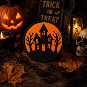 Könnte beinhalten: Ein kreisförmiger bestickter Aufnäher mit der schwarzen Silhouette eines Spukhauses und zwei kahlen Bäumen vor einem leuchtend orangefarbenen Hintergrund. Der Aufnäher hat einen dicken schwarzen Rand und liegt zwischen Halloween-Dekorationen.