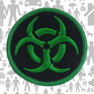 May include: A circular embroidered patch featuring a black background with a vibrant green biohazard symbol centered on the fabric. The patch is finished with a matching green stitched border that frames the circular design clearly.