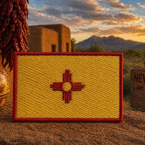 May include: A yellow and red New Mexico flag patch with the Zia symbol. The patch is on a textured surface, with a painted vase, dried red chiles, and a wooden sign that reads "New Mexico Land of Enchantment" in the background.