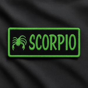 May include: A rectangular black patch with a green border and the word "SCORPIO" embroidered in green. A green scorpion graphic is on the left side of the patch. The patch is on a black fabric background.