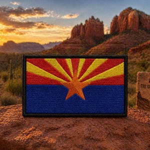 May include: Embroidered patch featuring the Arizona state flag. The flag design includes a blue lower section, a red and yellow sunburst, and a copper star. The patch is displayed on a rock with a desert landscape background.