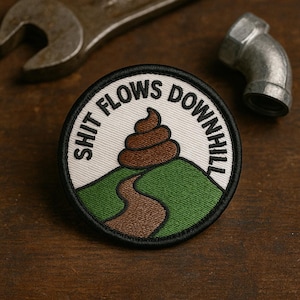 May include: Embroidered patch with the text "SHIT FLOWS DOWNHILL" around a brown poop emoji on a green hill with a brown path. The patch has a black border and is on a wooden surface.