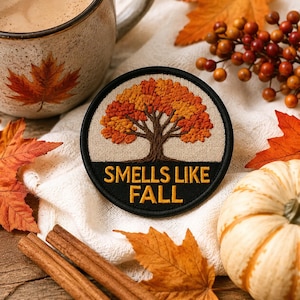 May include: A circular patch with a black border and the words "SMELLS LIKE FALL" in yellow. The patch features an embroidered tree with orange and brown leaves. The image includes a mug, autumn leaves, cinnamon sticks, and a small pumpkin.