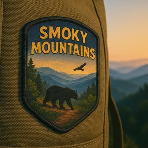 以下が含まれることがあります： 金色の文字で「SMOKY MOUNTAINS」と書かれたパッチ。パッチにはクマ、山、木、飛んでいる鳥が描かれています。パッチは緑色のバックパックに取り付けられています。