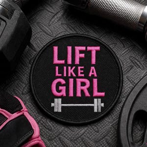 May include: A circular black embroidered patch featuring the pink text LIFT LIKE A GIRL and a silver barbell graphic. The patch has a stitched border and is displayed among gym equipment including a dumbbell, weight plate, and pink gloves.