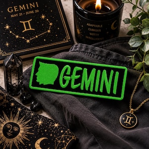 Op de afbeelding: Een rechthoekige geborduurde patch met een felgroen silhouet van een profiel en het woord GEMINI in een bijpassend groen lettertype op een zwarte achtergrond. De patch ligt op een donkere stoffen ondergrond naast andere astrologische items.