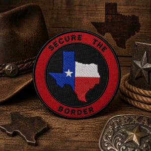 May include: A round patch with the Texas state flag design, featuring a blue star and the state outline in red, white, and blue. The text "SECURE THE BORDER" is in red around the edge. The patch is on a wooden surface with a cowboy hat and belt buckle.