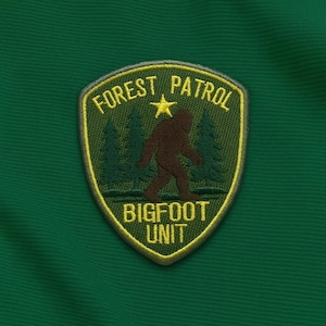 Puede incluir: Parche bordado con diseño de escudo. El parche presenta el texto "FOREST PATROL" y "BIGFOOT UNIT" en amarillo, con una silueta marrón de Bigfoot y árboles verdes. El parche está sobre un fondo verde.