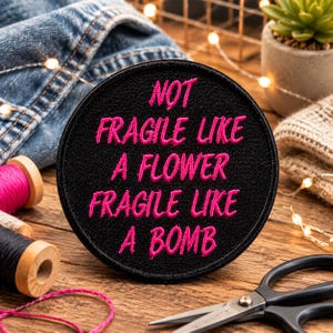 Op de afbeelding: Een ronde, zwarte geborduurde patch met de tekst "NOT FRAGILE LIKE A FLOWER FRAGILE LIKE A BOMB" in felroze letters. De patch ligt op een houten oppervlak met naaigerei en denim op de achtergrond.
