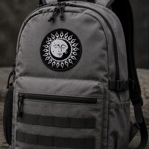 May include: A charcoal grey backpack featuring a circular embroidered patch of a sun with a human face. The bag includes a front zippered pocket, two horizontal webbing straps, side mesh pockets, black zips, and adjustable shoulder straps.