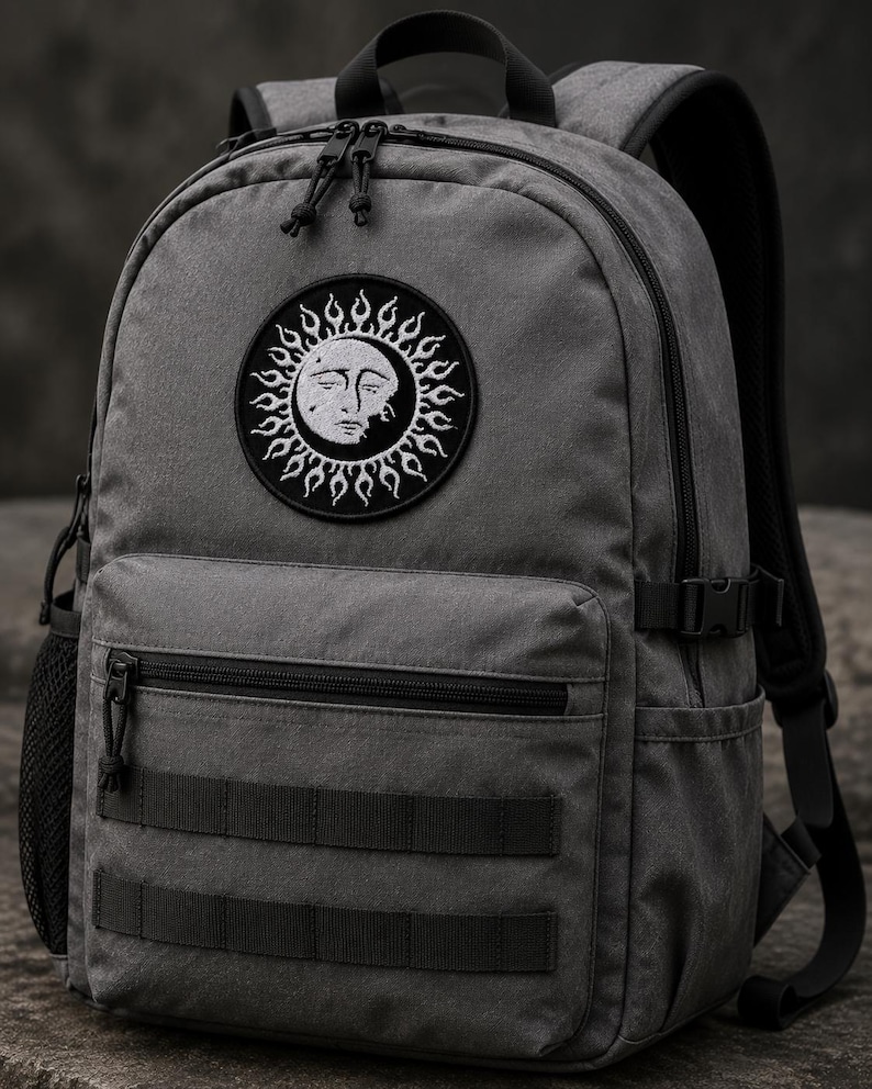 May include: A charcoal grey backpack featuring a circular embroidered patch of a sun with a human face. The bag includes a front zippered pocket, two horizontal webbing straps, side mesh pockets, black zips, and adjustable shoulder straps.