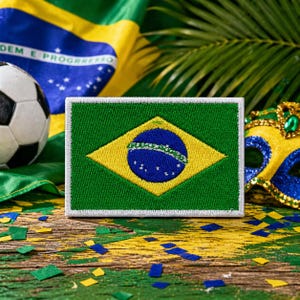 Puede incluir: Parche bordado con la bandera brasileña, con fondo verde, rombo amarillo y círculo azul. Un balón de fútbol, la bandera brasileña y una máscara decorativa están en el fondo. El parche está sobre una superficie de madera.