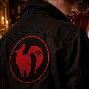 Pode incluir: Um casaco preto com um grande emblema circular nas costas. O emblema apresenta a silhueta de uma raposa bordada a vermelho com um s&iacute;mbolo solar na cauda, rodeado por um rebordo circular fino e vermelho sobre um fundo preto.