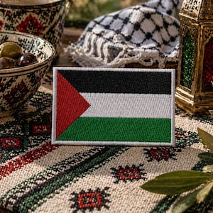 Puede incluir: Parche rectangular bordado con la bandera de Palestina. El diseño de la bandera incluye franjas horizontales negras, blancas y verdes, y un triángulo rojo. El parche está sobre una superficie estampada, con aceitunas en un cuenco y una linterna decorativa.